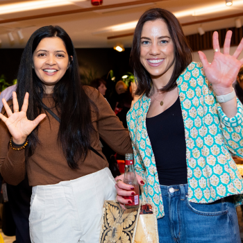 Two smiling women wave at the camera while attending a social event, holding 5 fingers up, with a lively crowd in the background.